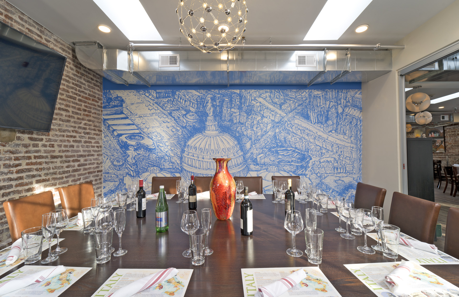 Long dining table set with glasses, plates, and napkins in a modern room with a blue mural wall and chandelier.