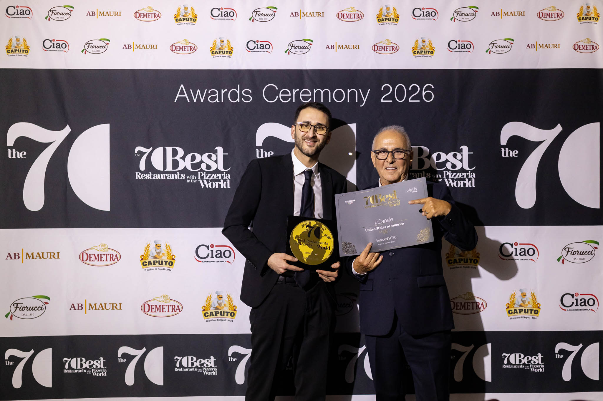 Two men in formal suits holding awards and a certificate at an awards ceremony backdrop.