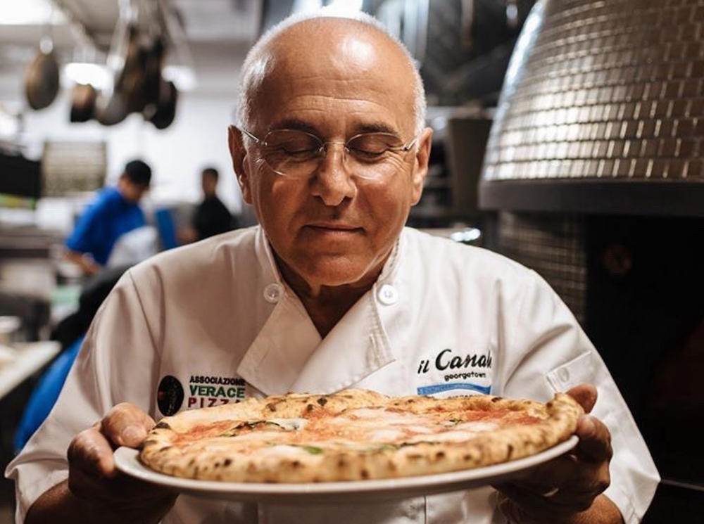 Chef holding a pizza in a kitchen with blurred background of people and kitchen equipment.
