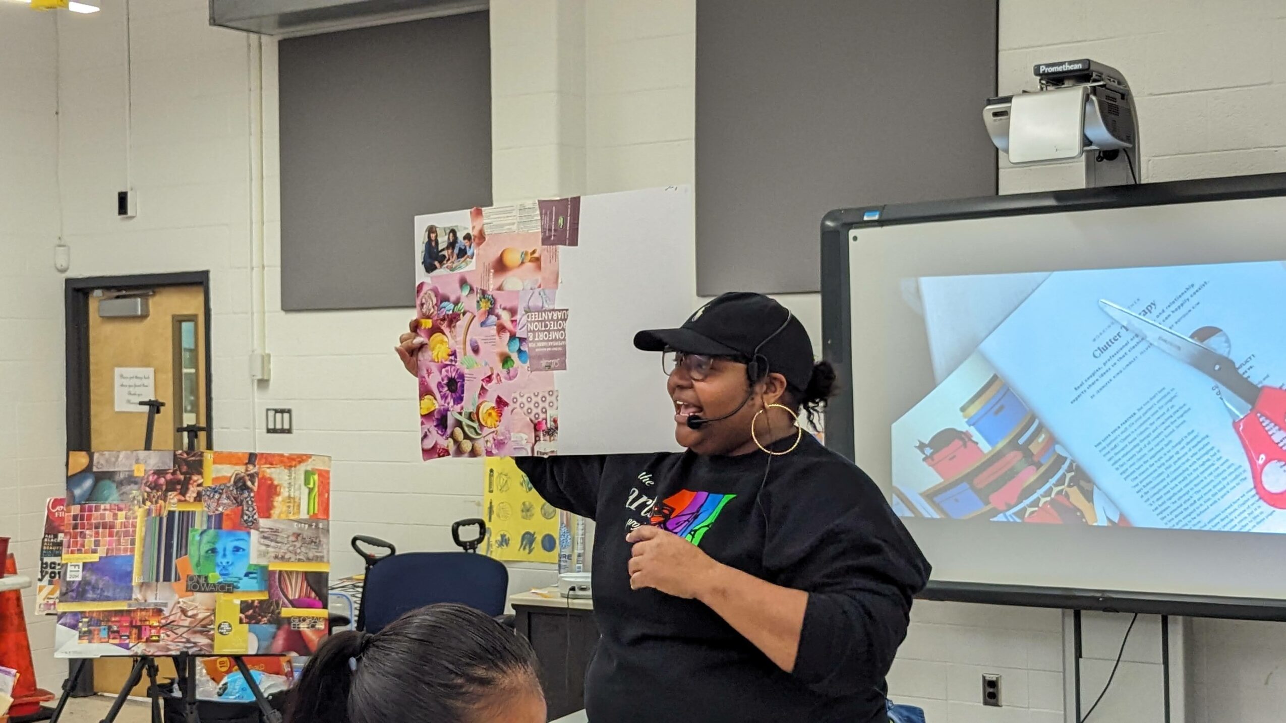 Person wearing a black cap and glasses speaking in front of a classroom with colorful artwork and a large screen.
