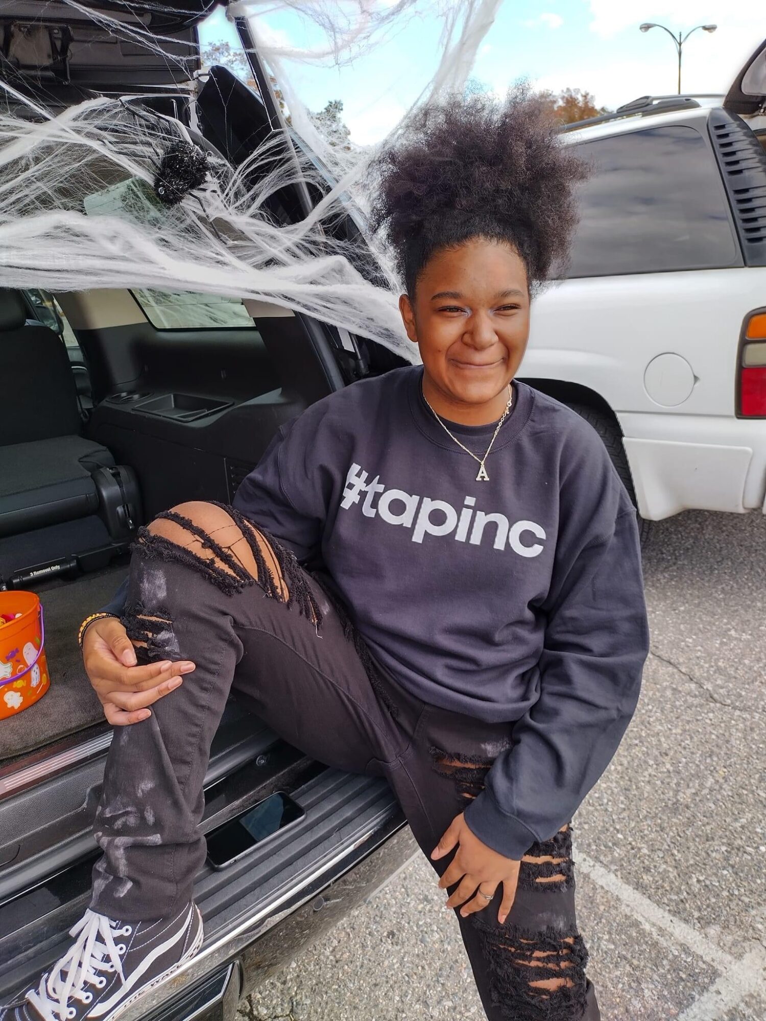 Girl with curly hair smiling, sitting on car door, wearing a black hoodie with #apinc, ripped jeans, and sneakers.