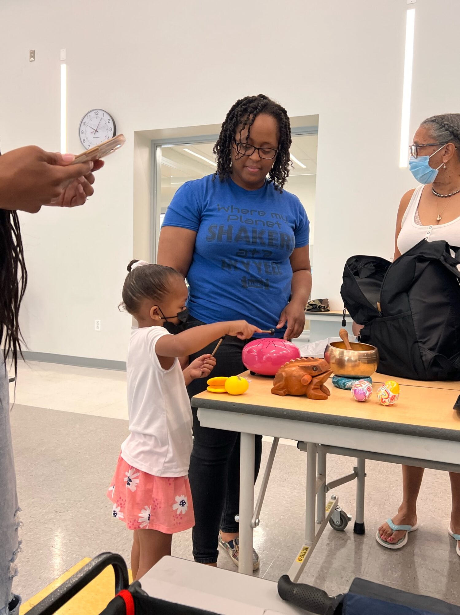 Child and woman looking at items on table, woman wearing face mask, person holding phone nearby, plain room background.