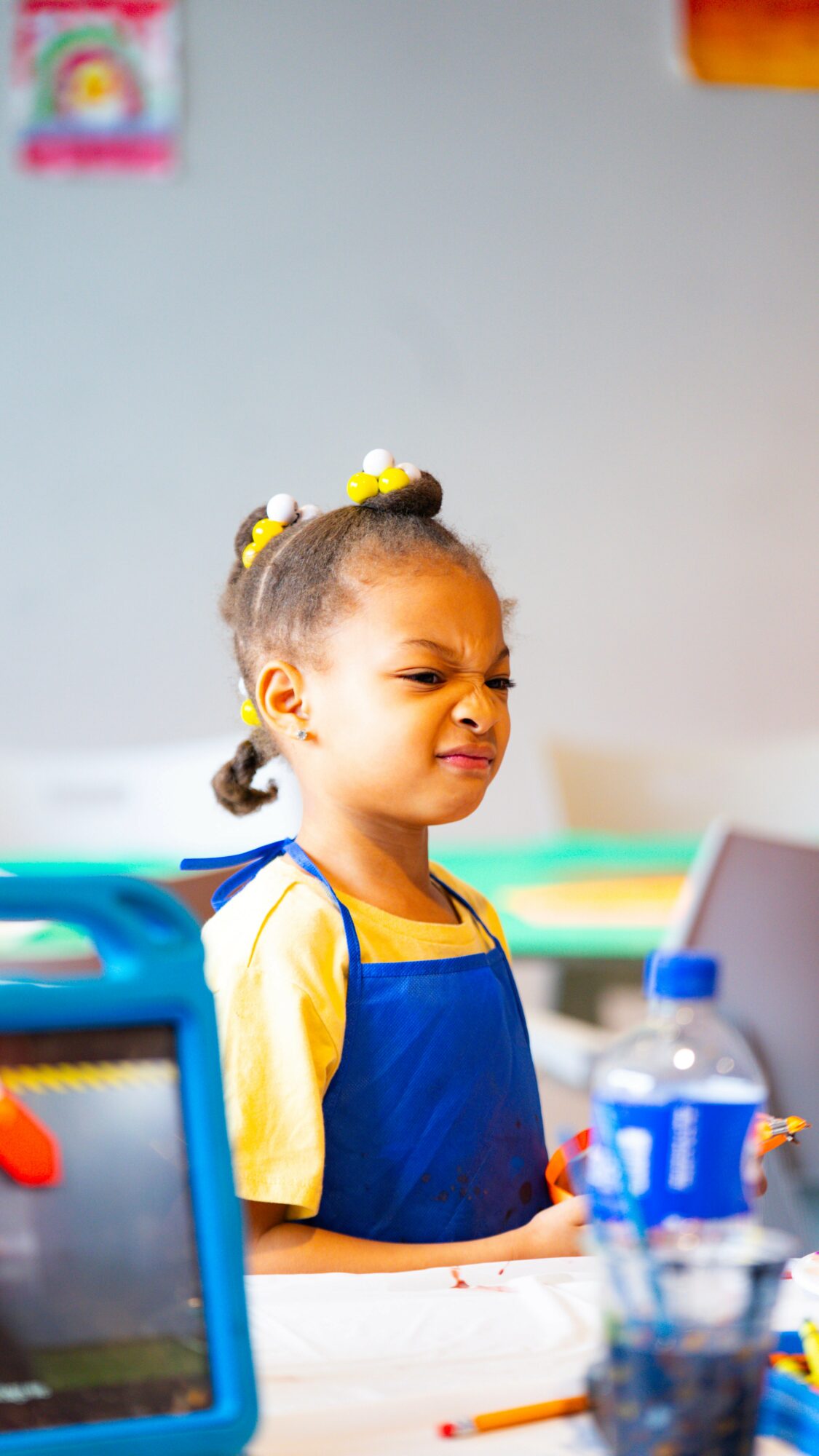 Young girl with braided hair and yellow hair accessories, wearing a blue apron, in a classroom or activity setting.