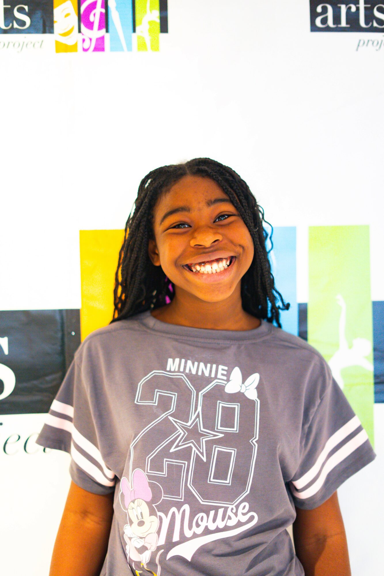 Young girl with long curly hair smiling, wearing a Minnie Mouse T-shirt, standing in front of a colorful background.