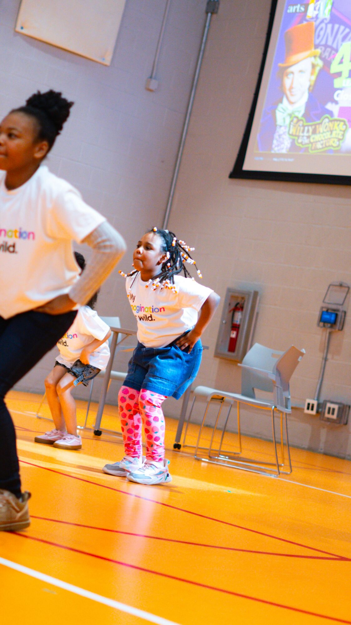 Children dancing in a gymnasium with a screen displaying a cartoon character in the background.