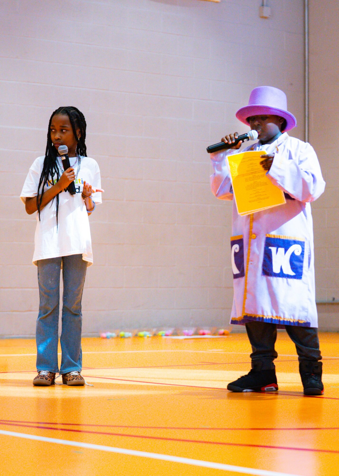Two children on stage, one holding a microphone and the other reading from a paper, in a gymnasium.