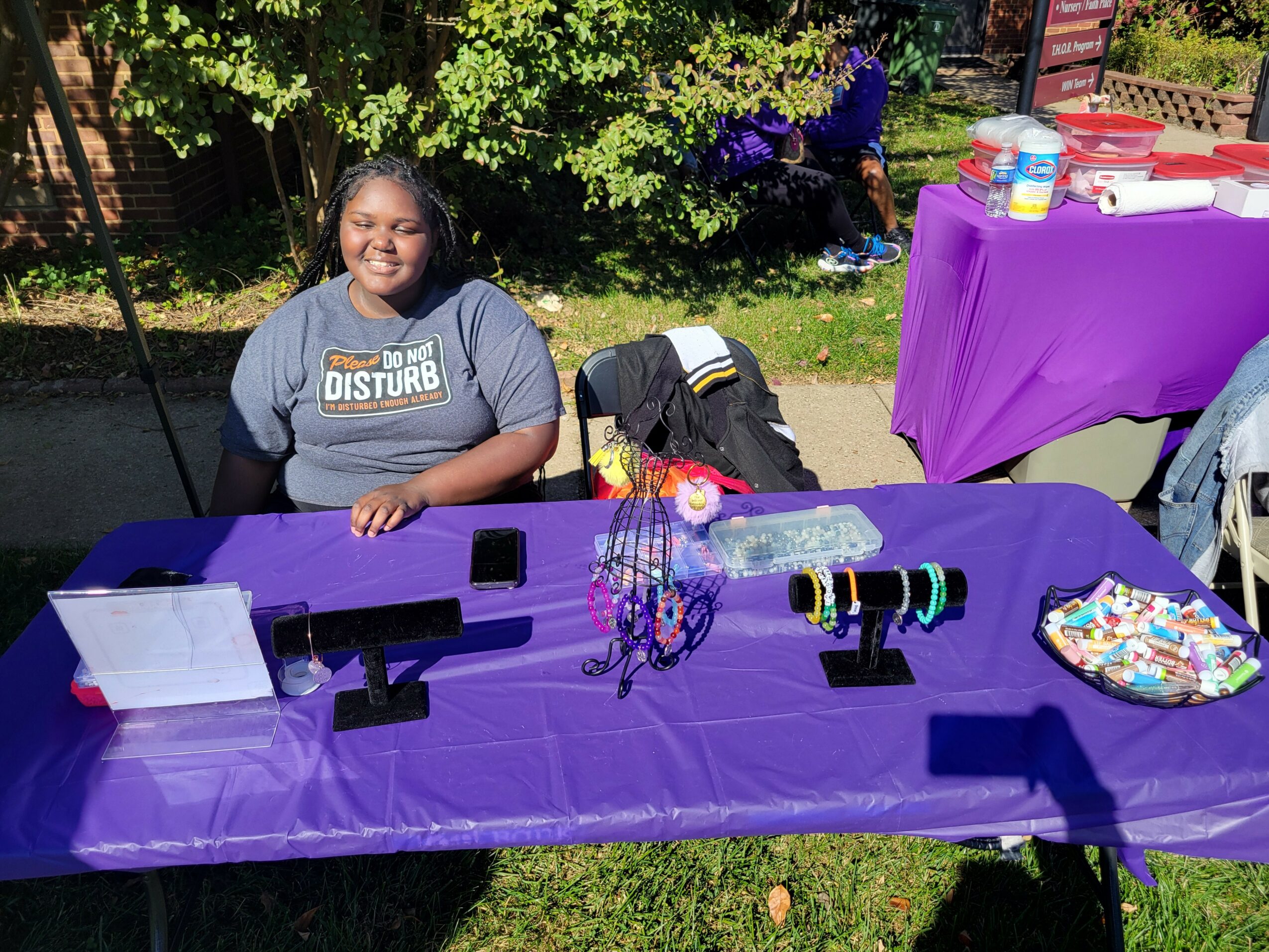 Smiling girl sitting behind a purple table outdoors with colorful items and a purple tablecloth.