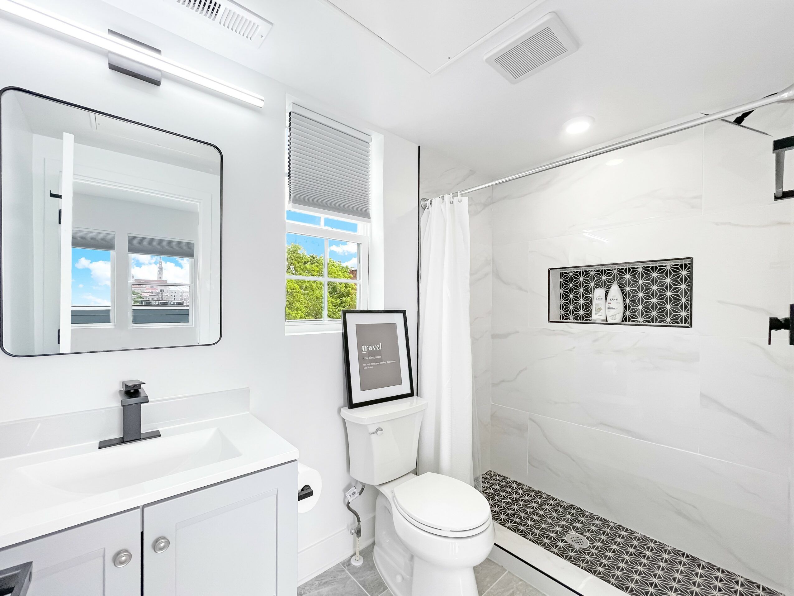Bathroom with white walls, vanity, mirror, toilet, window, and shower area with black and white tiles.