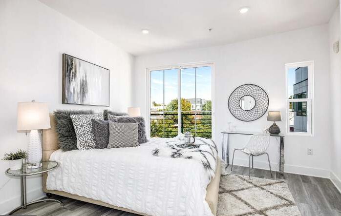 Bright bedroom with white walls, large window, bed with gray and white pillows, and a desk with a chair.