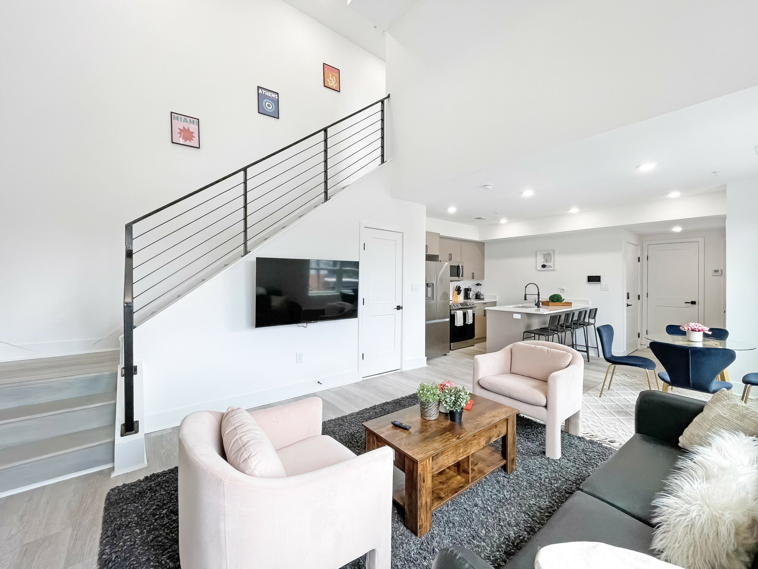 Living room with two armchairs, a sofa, a wooden coffee table, and a TV on the wall, with stairs and kitchen in background.