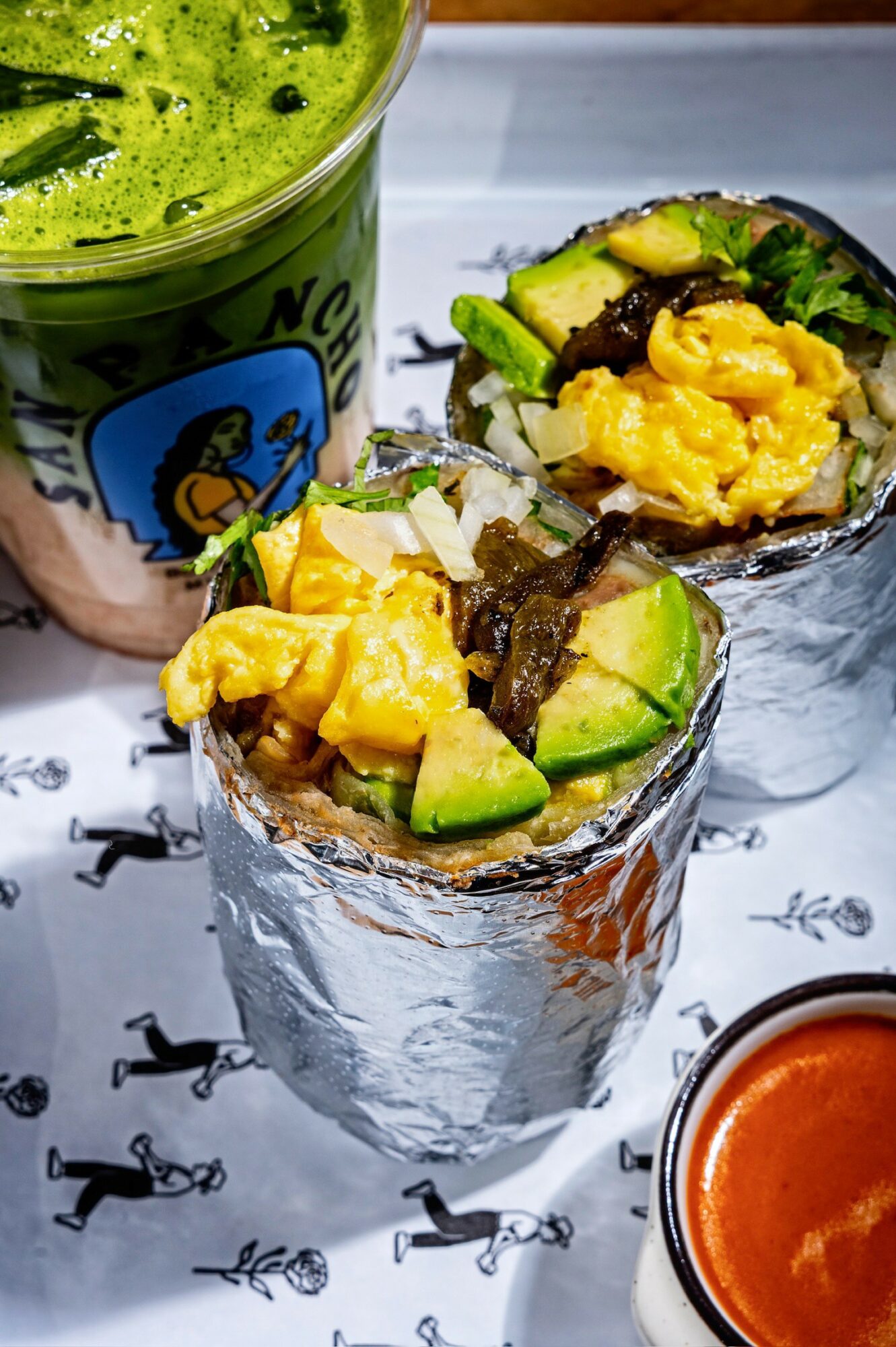 Two foil-wrapped avocado and scrambled egg tacos on a white surface with a green drink and red sauce.