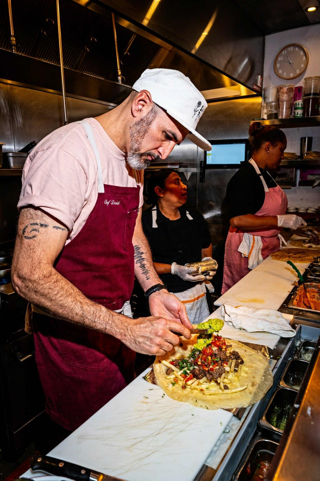 Man in white cap and apron decorating a cake in a kitchen with two women in the background.
