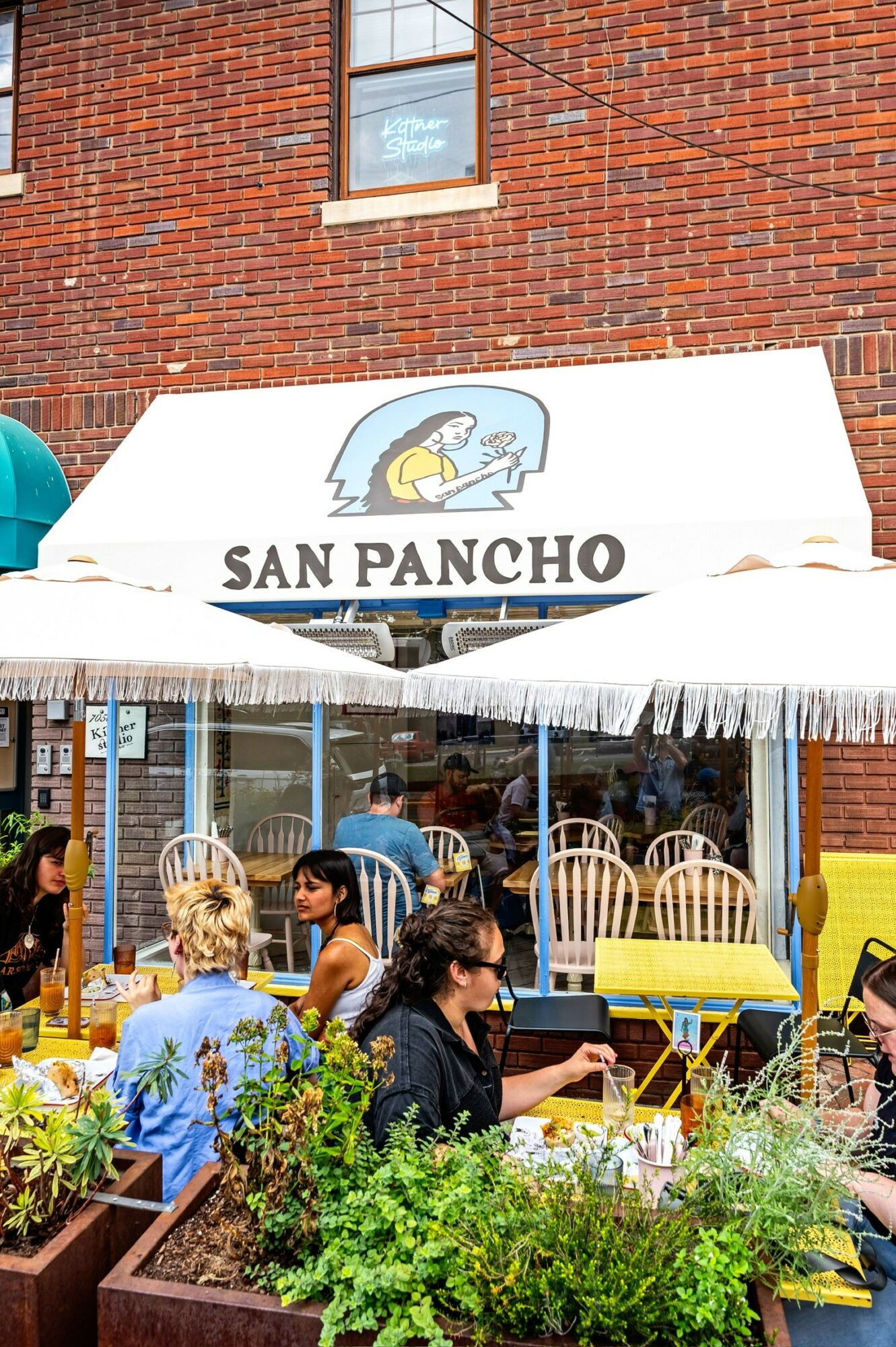 Outdoor seating area with people dining, plants, and a white awning with a sign for San Pancho, brick building background.