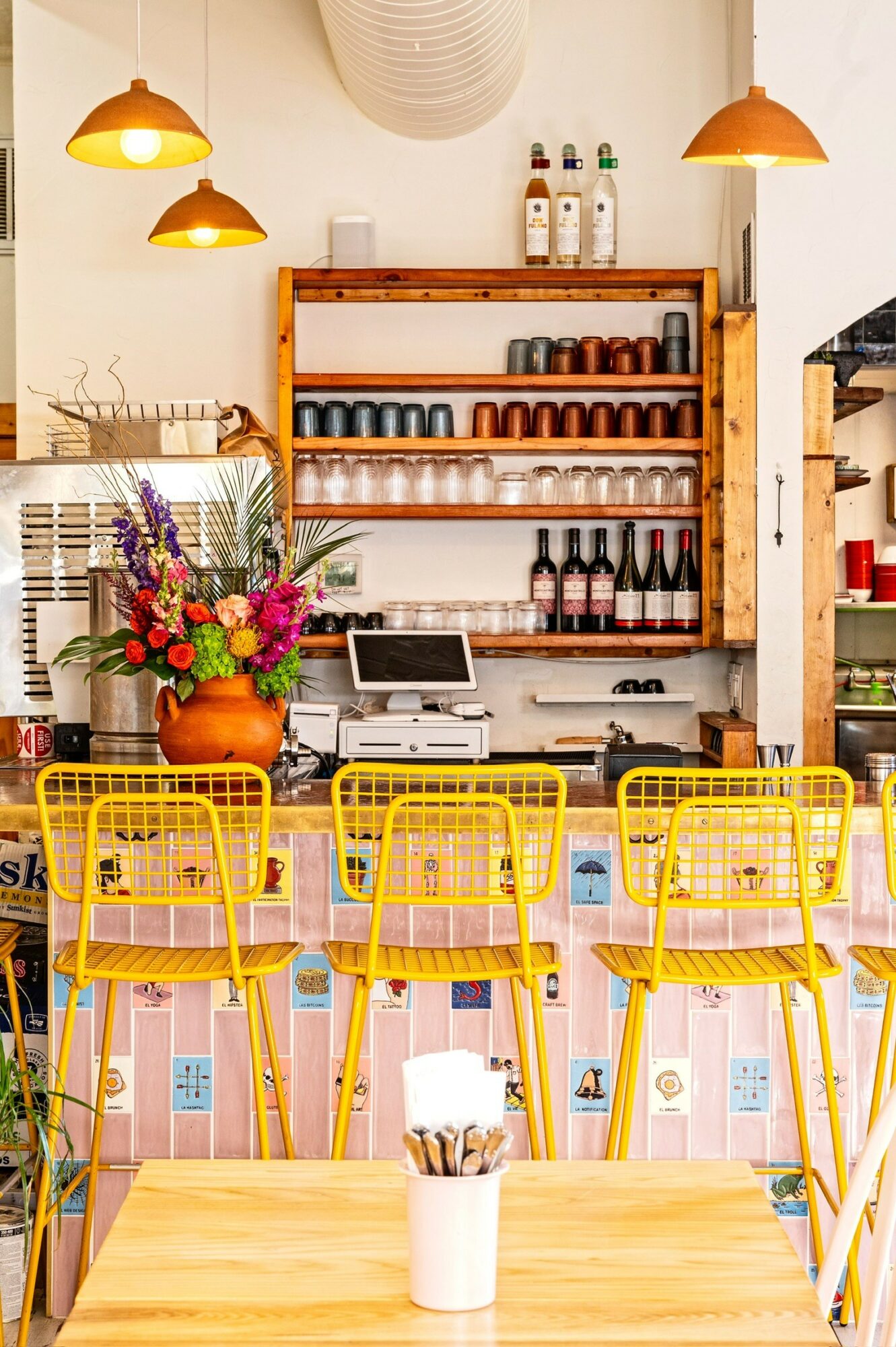 Interior of a cafe with yellow chairs, a wooden table, and shelves with bottles and cups.