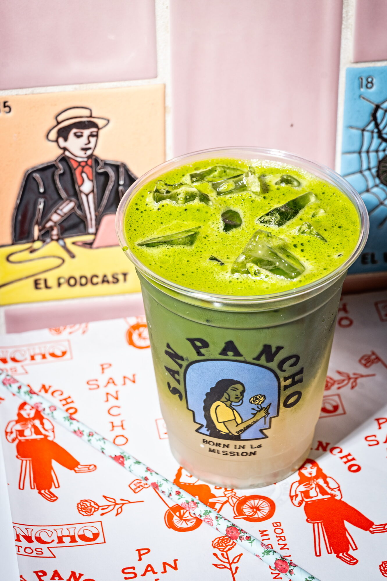 Green beverage with ice in a cup featuring a cartoon character and text, with colorful posters and drawings in background.