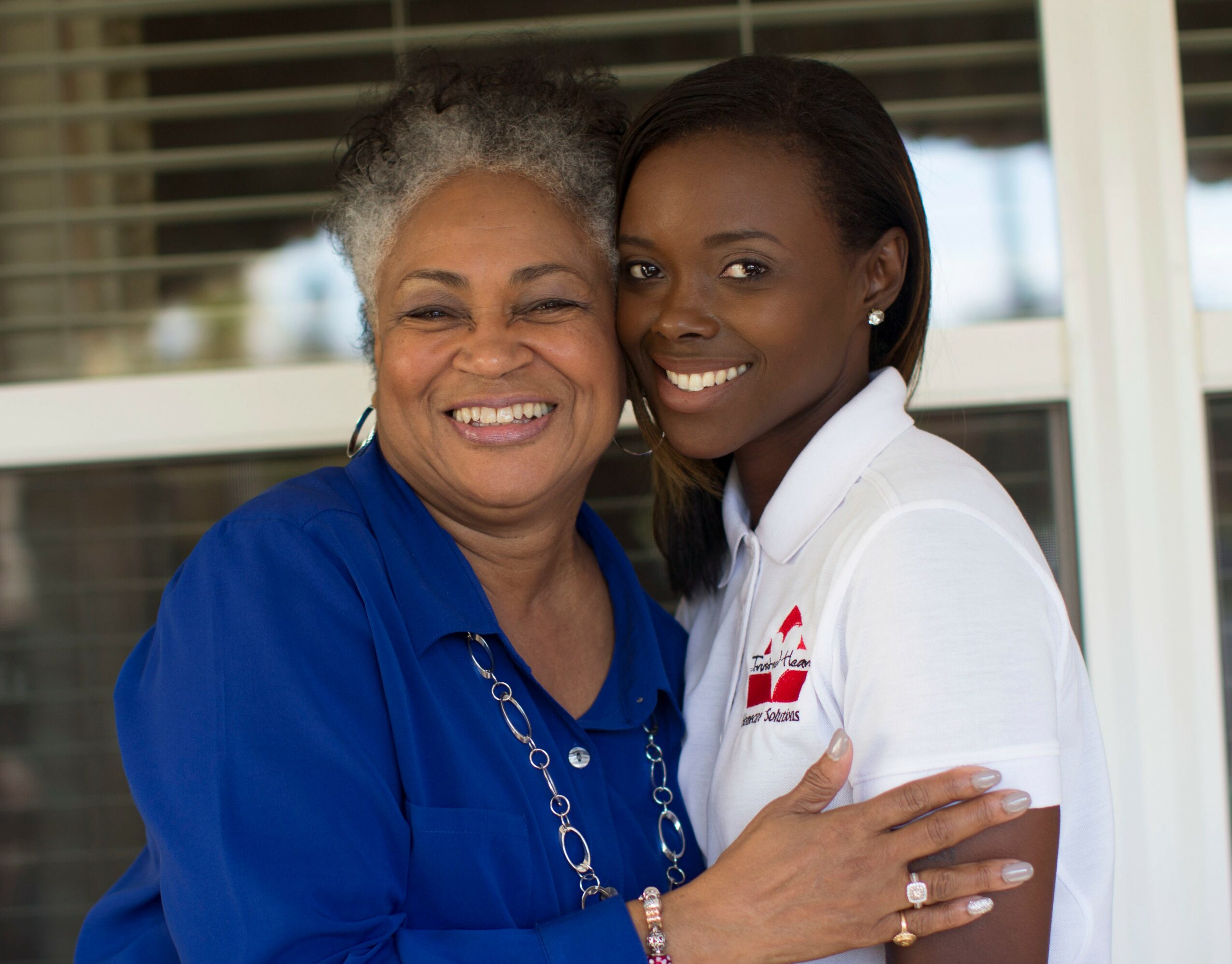 Two women smiling and hugging outdoors, one older with gray hair, the other younger with dark hair, both in casual clothing.