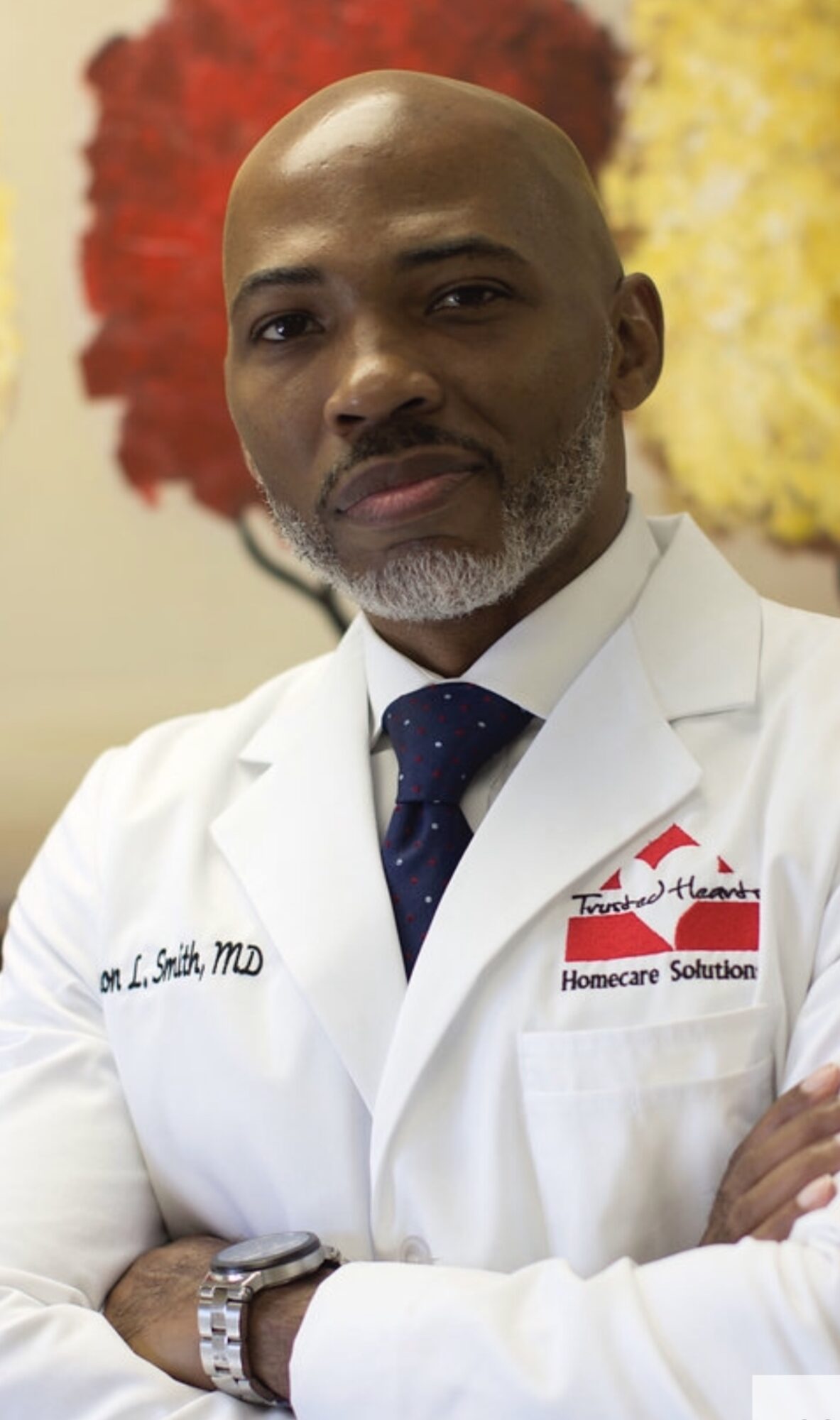 Man in white coat with a blue tie, standing with arms crossed, background of colorful flowers.