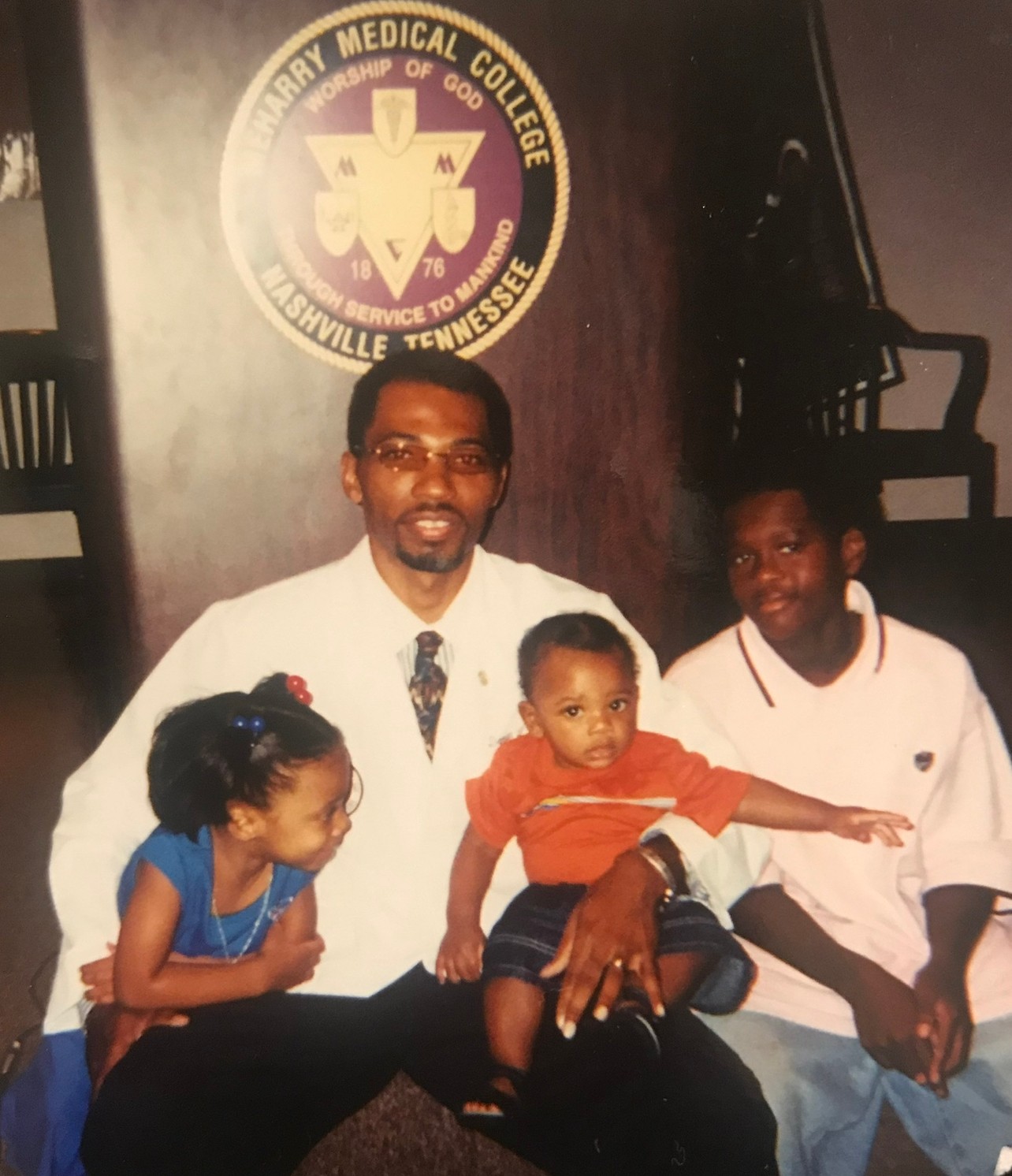 Man with glasses and beard holding a child, two children beside him, in front of a Tennessee medical college sign.