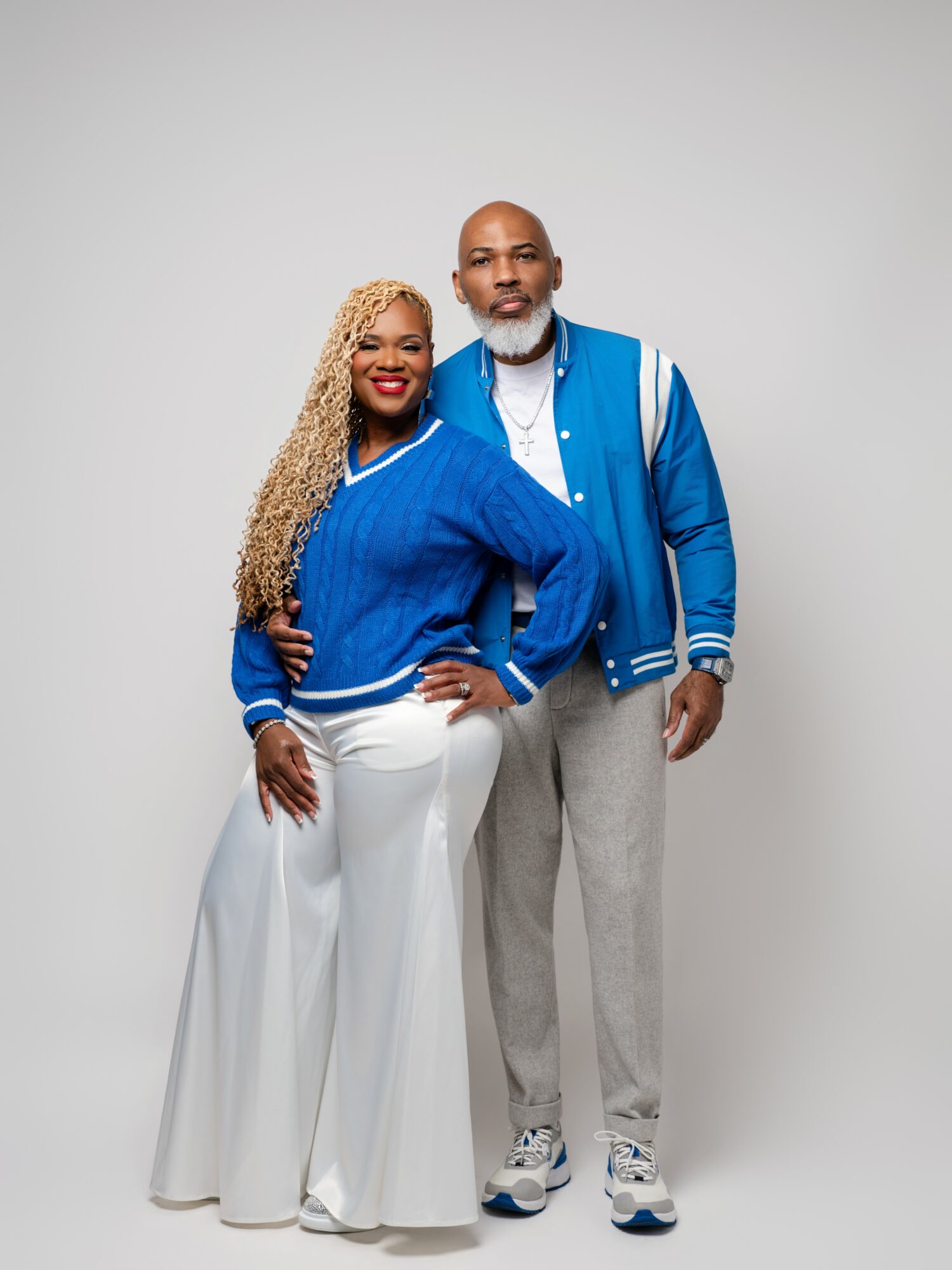 A smiling couple stands together against a plain background, with the woman wearing a blue jacket and white pants, and the man in a blue jacket and gray pants.