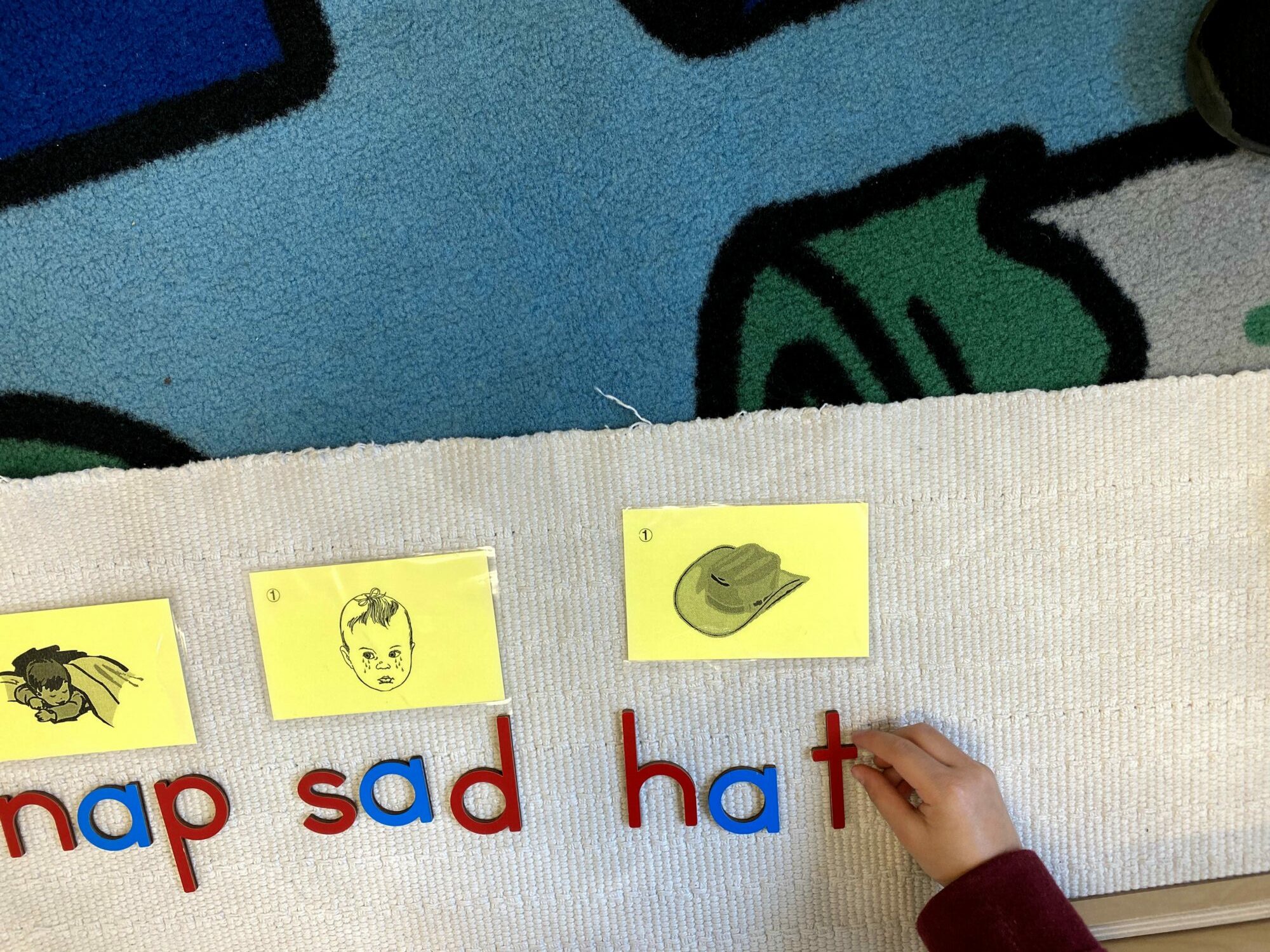 Child's hand points to a yellow card with a drawing of a hat, on a classroom rug with letters and images.