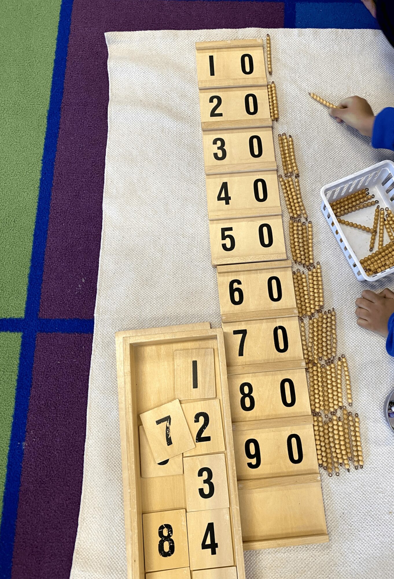 Educational number tiles arranged vertically with a person using a pen nearby, on a table with colorful fabric.