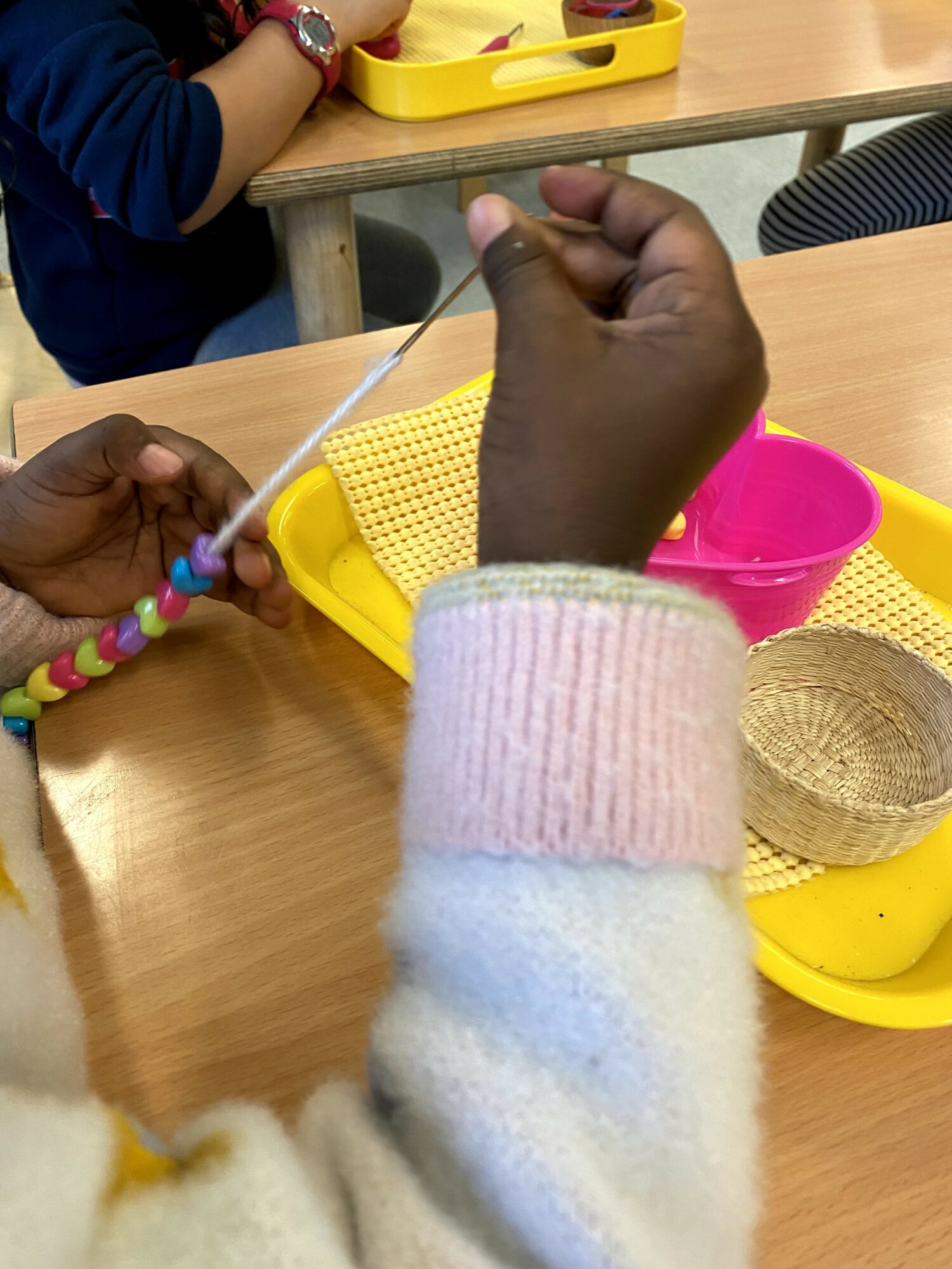 Child's hands holding colorful beads and string, with yellow trays and pink bowl on table.