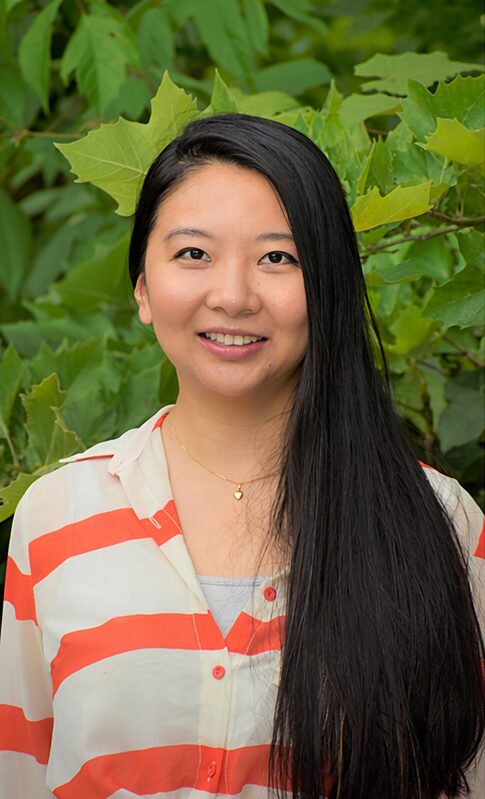 Young woman with long black hair smiling outdoors, surrounded by green leaves, wearing a striped shirt and a necklace.