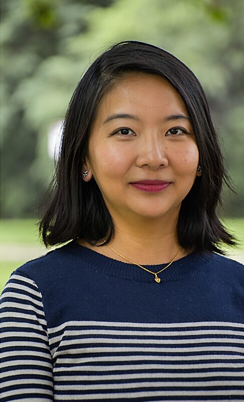 Portrait of a woman with shoulder-length black hair, wearing a navy and white striped top, outdoors with green background.