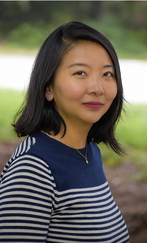 Young woman with shoulder-length black hair, wearing a striped navy and white top, outdoors with greenery background.
