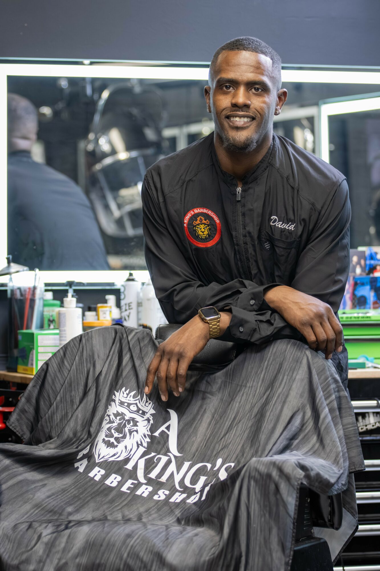 Man with short hair and beard smiling, wearing a black jacket, sitting in a barbershop with tools and products nearby.