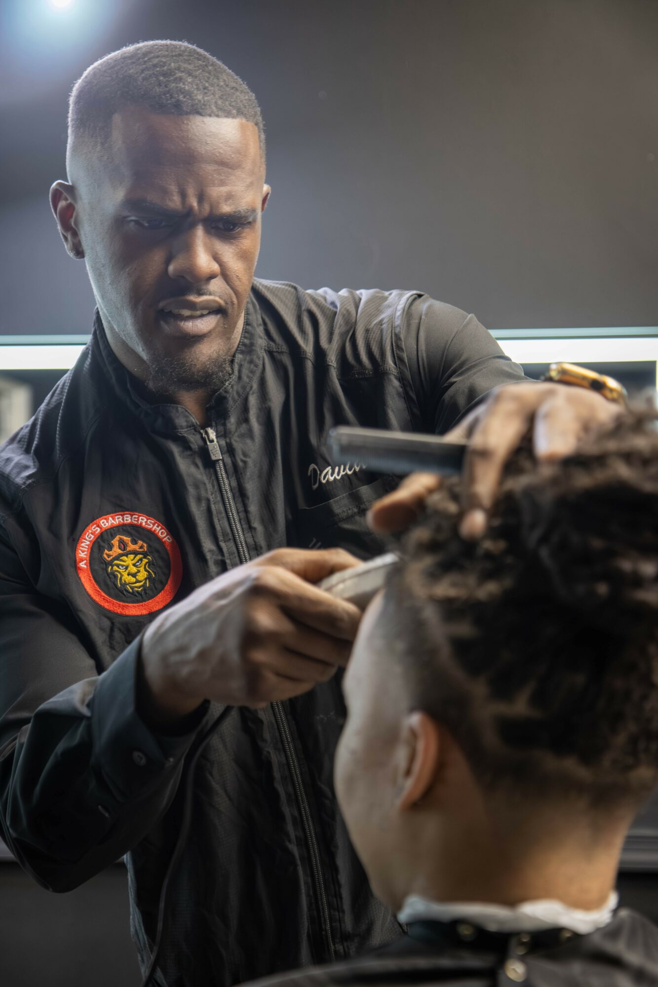 Barber cutting a person's hair in a salon, focused expression, dark jacket with a patch, hair styling tools visible.