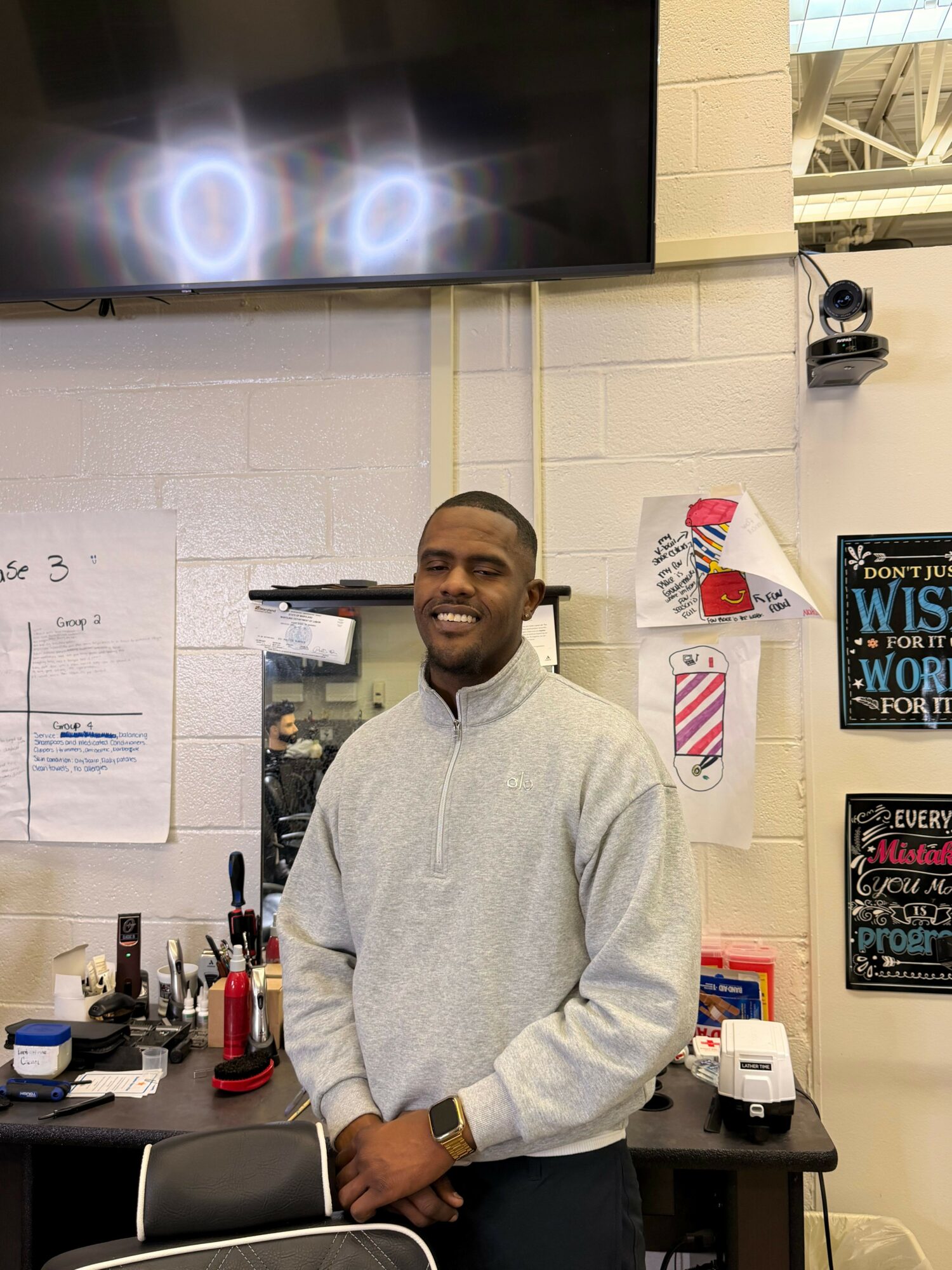 Young man smiling, standing in a classroom or office, wearing a light gray sweatshirt, with various posters and objects behind him.