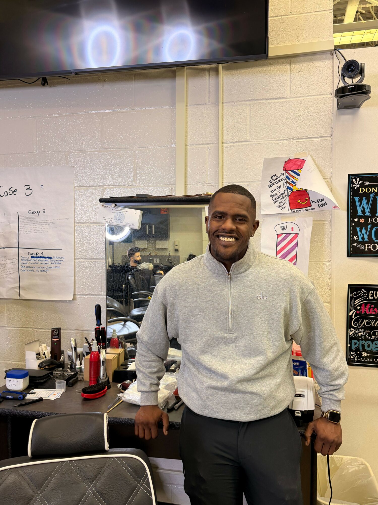 Smiling man in gray sweater standing in a room with various objects and posters on the wall.
