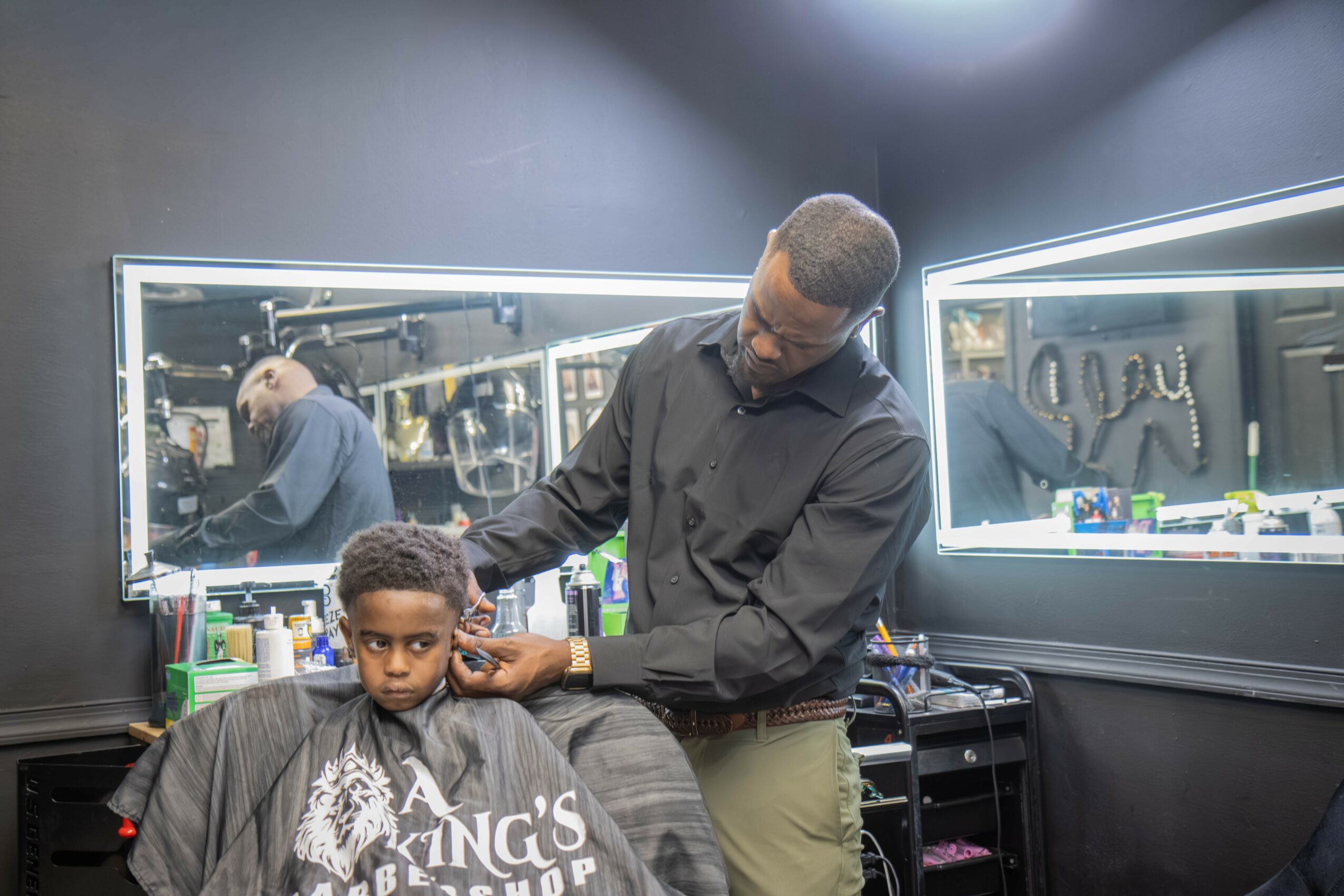 Barber cutting a child's hair in a barbershop with mirrors and tools visible.