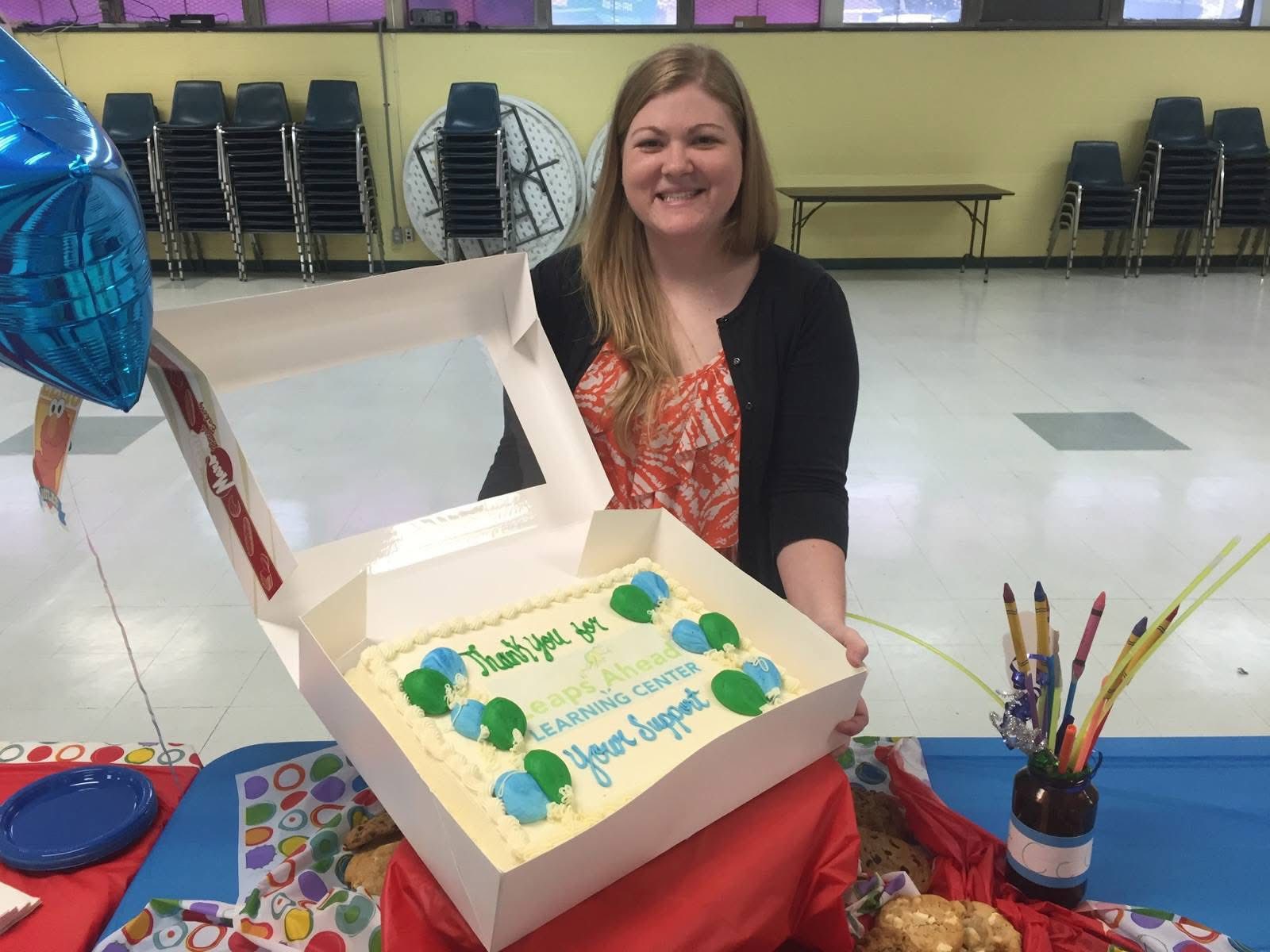 Smiling woman sitting behind a decorated cake with green and blue decorations, in a room with chairs and tables in the background.