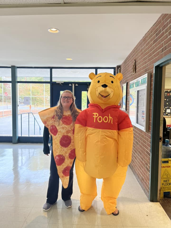 Person standing next to a Winnie the Pooh mascot costume in a hallway with glass doors and brick wall.
