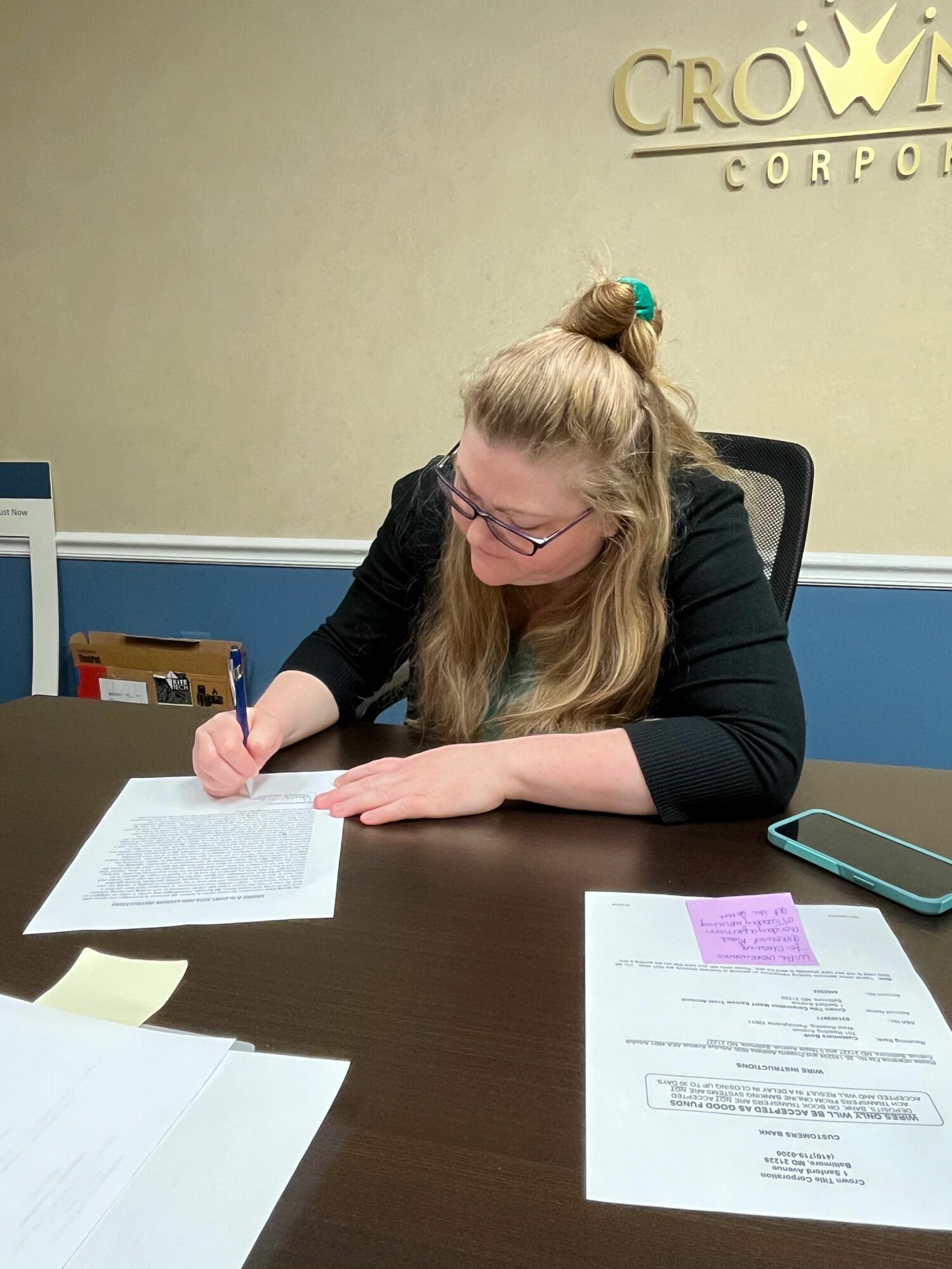 Woman with glasses and long hair tied back, writing on paper at desk in office.