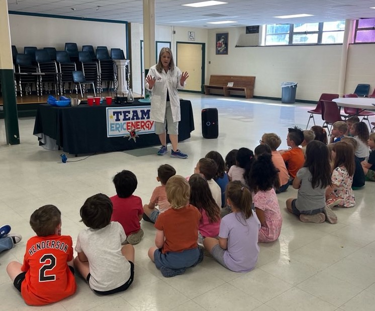 Woman in white coat speaking to children seated on floor in a large room with tables and chairs.