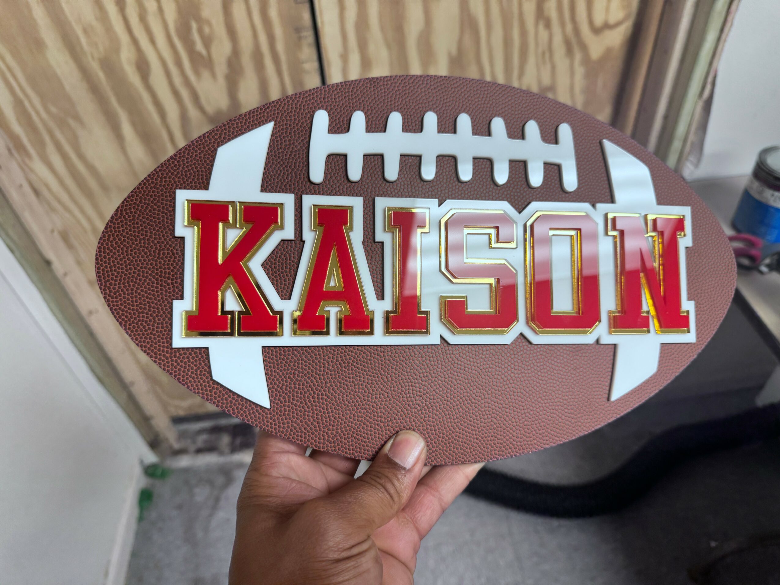 Hand holding an oval football-shaped sign with the name 'KAISON' and a football graphic.