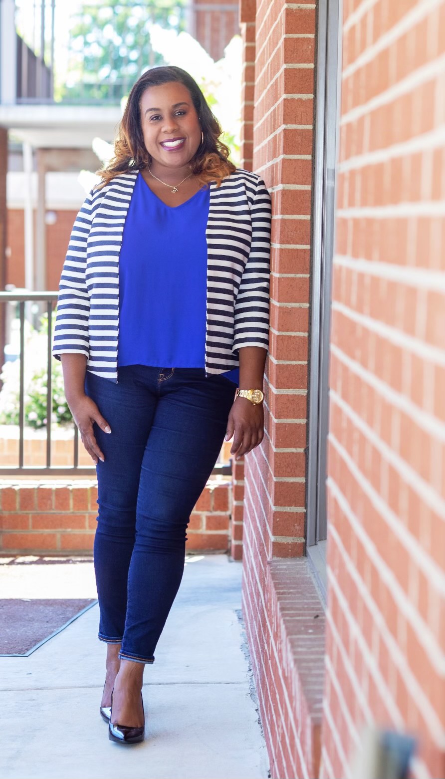 Woman standing outdoors near brick wall, smiling, wearing blue top, striped jacket, jeans, and heels.