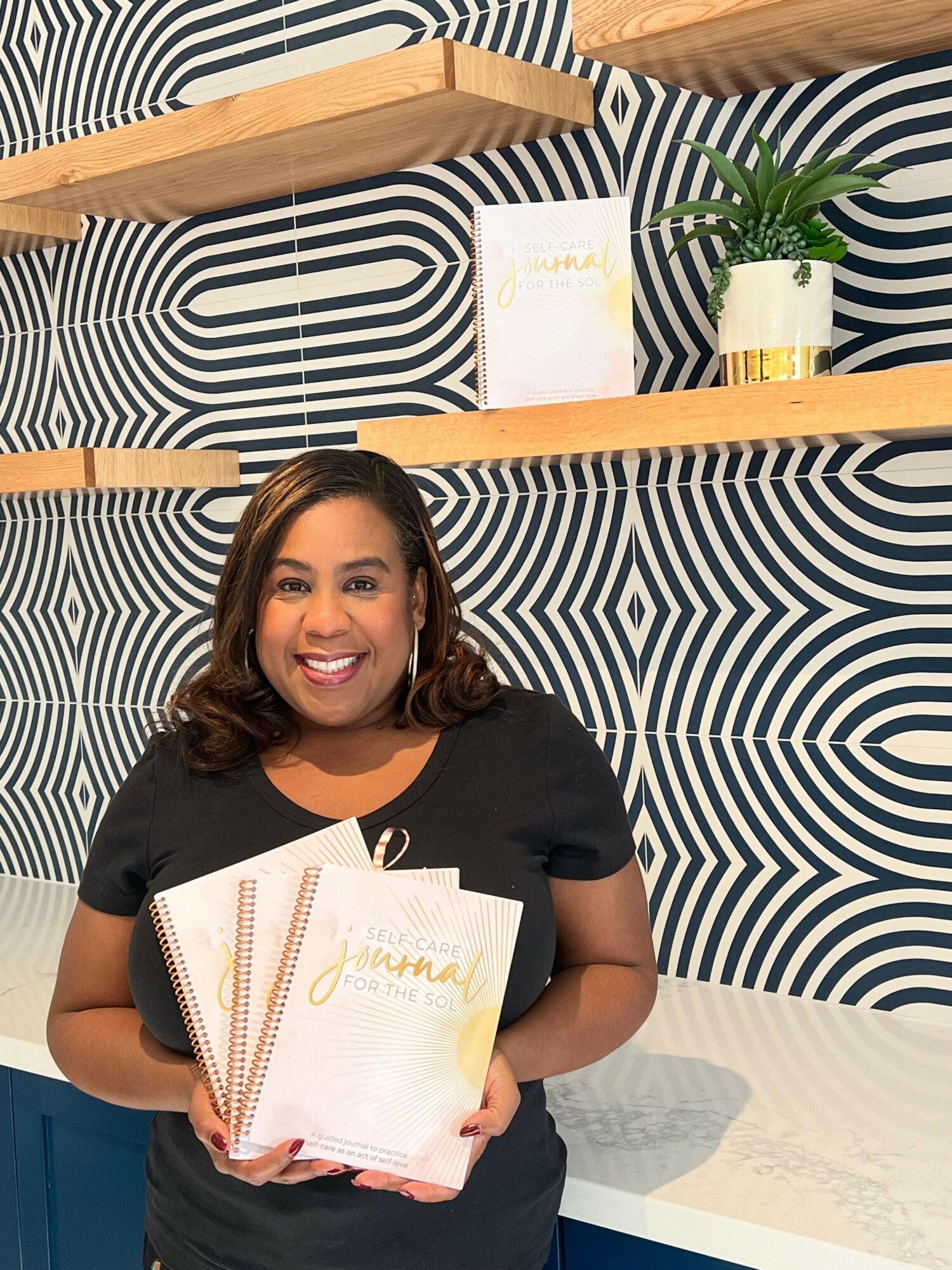 Smiling woman holding notebooks in a room with patterned wall and wooden shelves.
