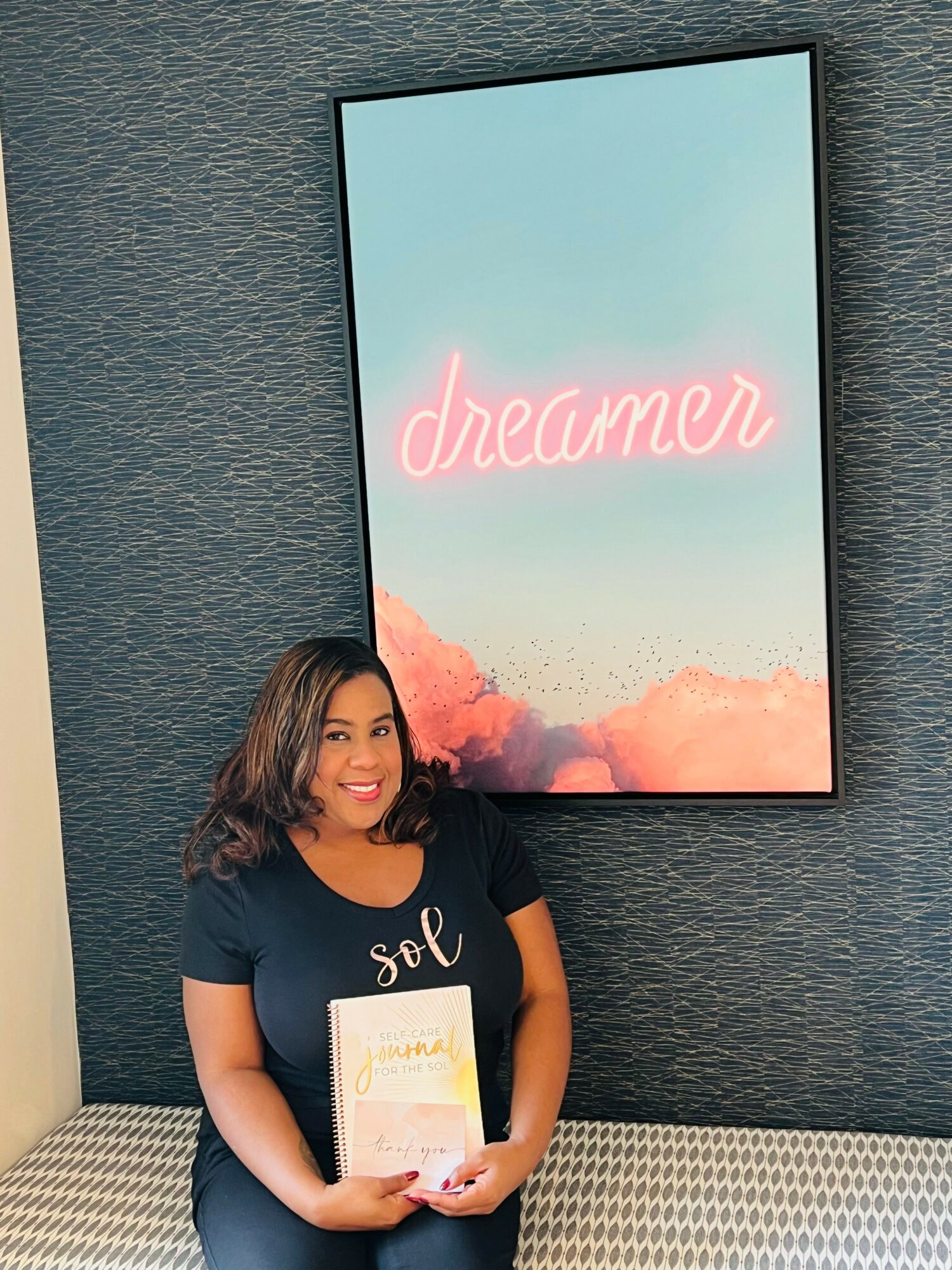 Smiling woman holding a book in front of a textured wall with a large screen displaying the word 'dreamer' in pink neon.