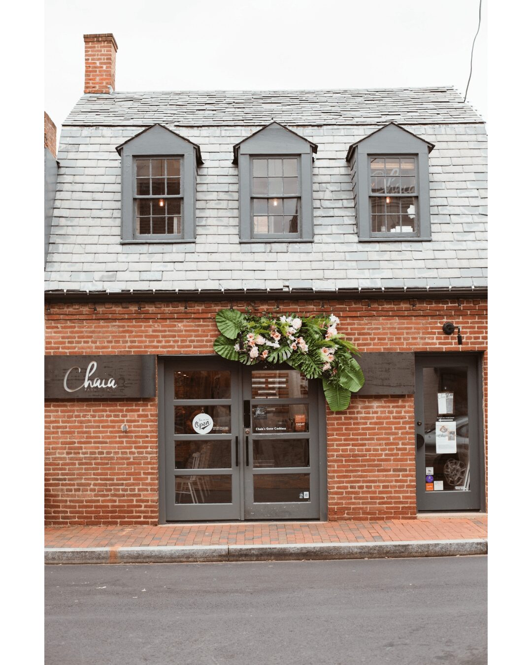 Brick building with three dormer windows on gray shingled roof and a door with a plant decoration above.