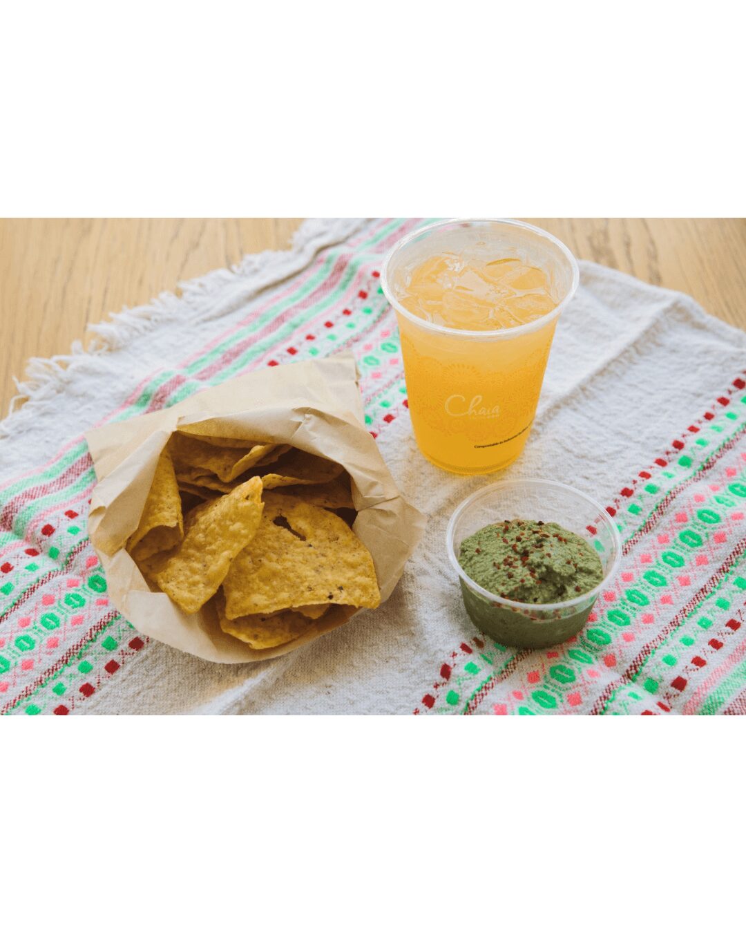 Bag of tortilla chips, a cup of orange drink, and a small bowl of green salsa on a cloth-covered table.