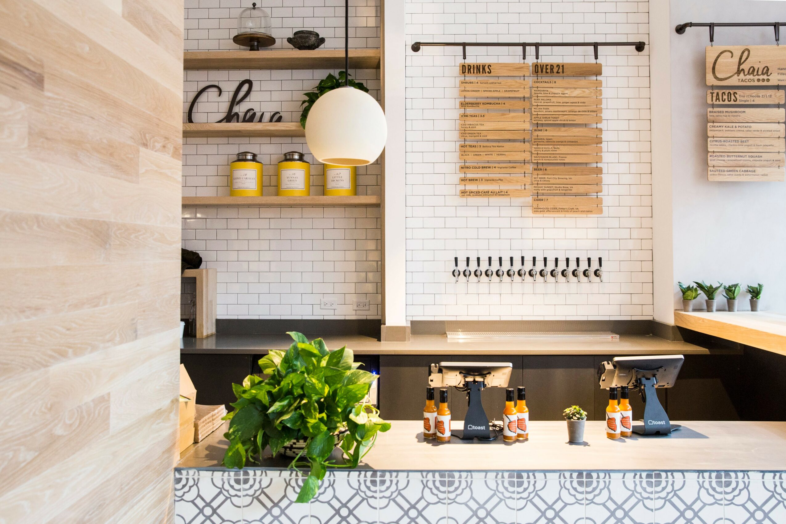 Kitchen with white tiled wall, wooden shelves, potted plants, and a counter with bottles and a plant.