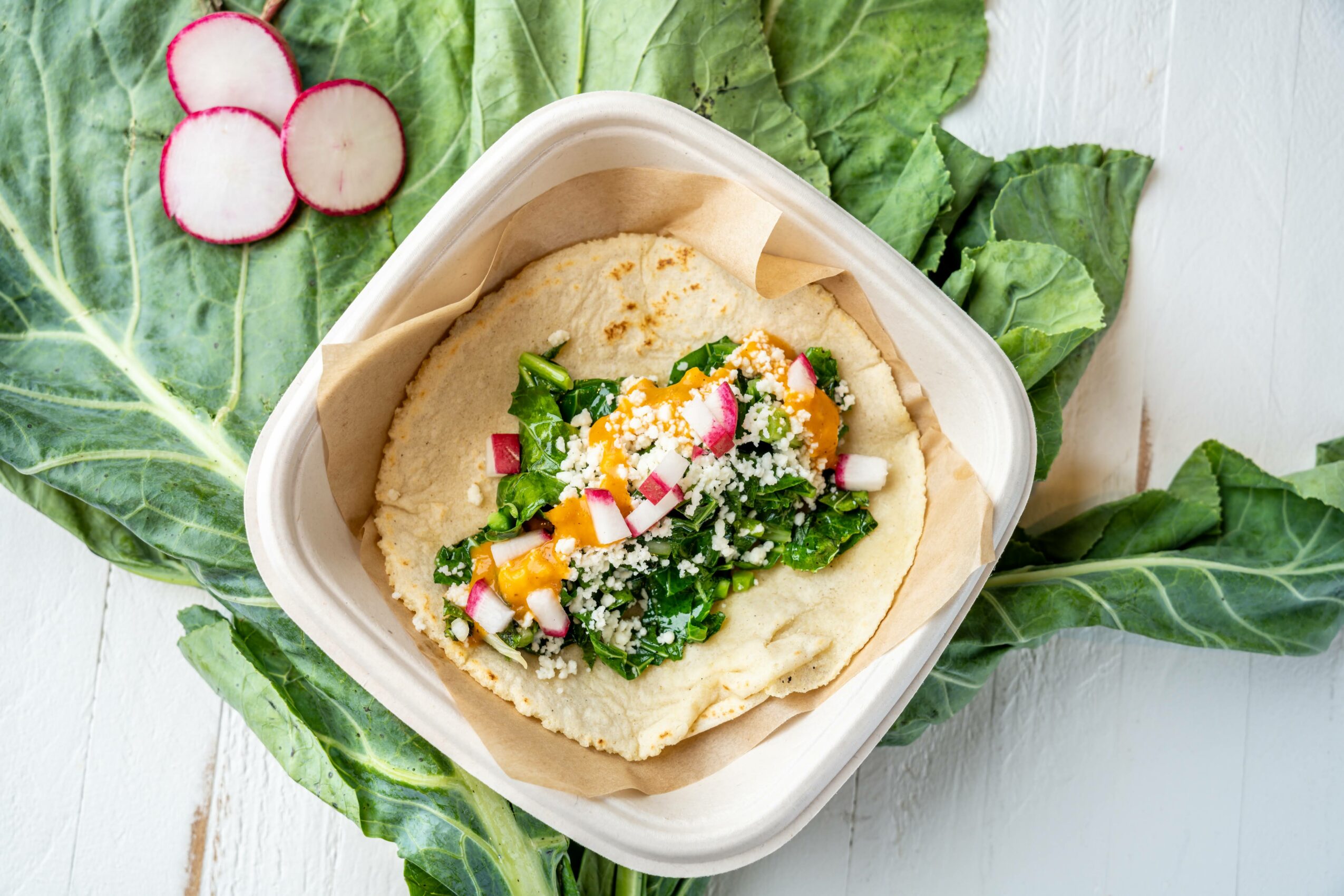 Bowl of hummus topped with chopped vegetables and herbs, surrounded by leafy greens and radishes on a white surface.