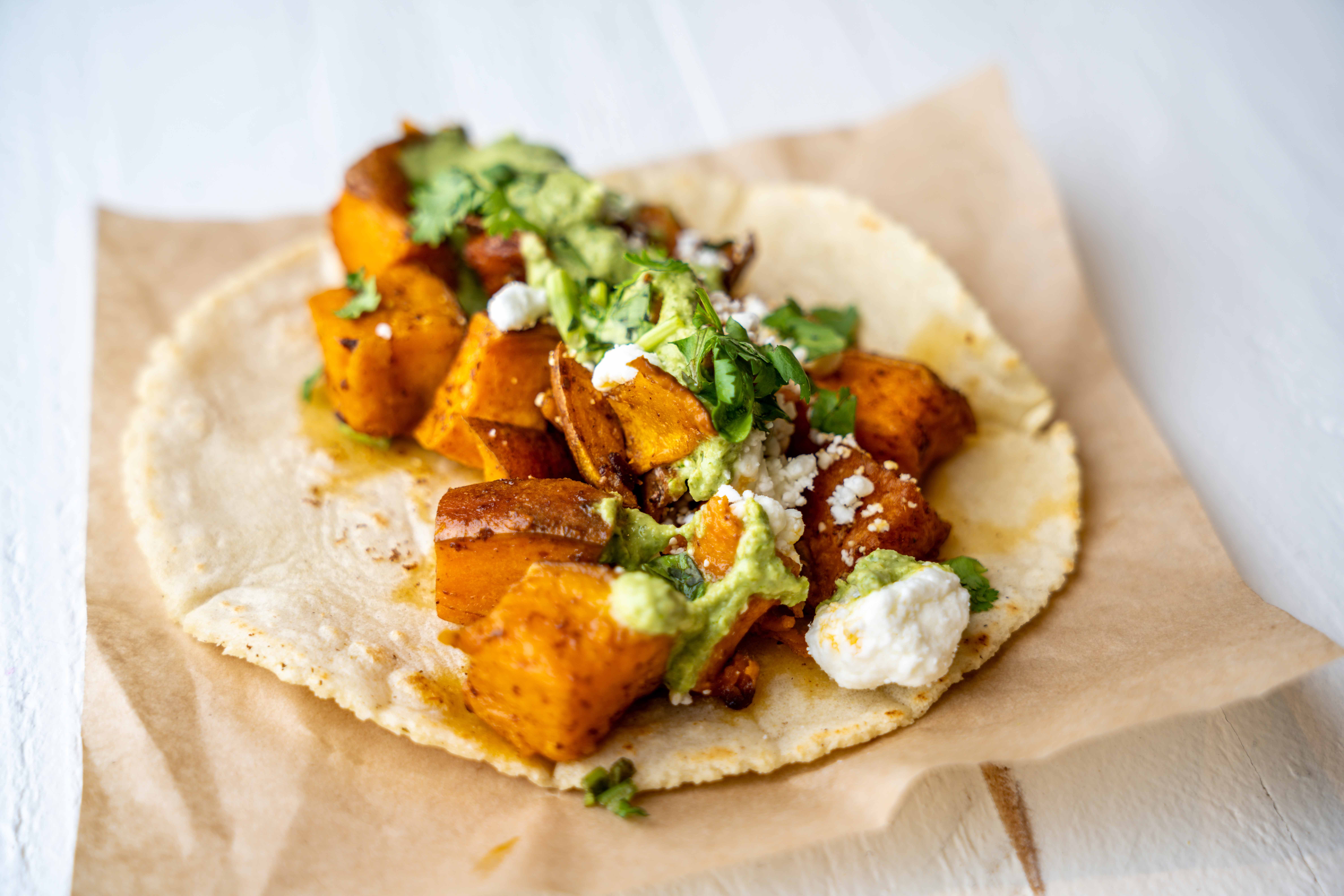 Taco with roasted sweet potatoes, avocado, cheese, and herbs on a paper-lined surface.
