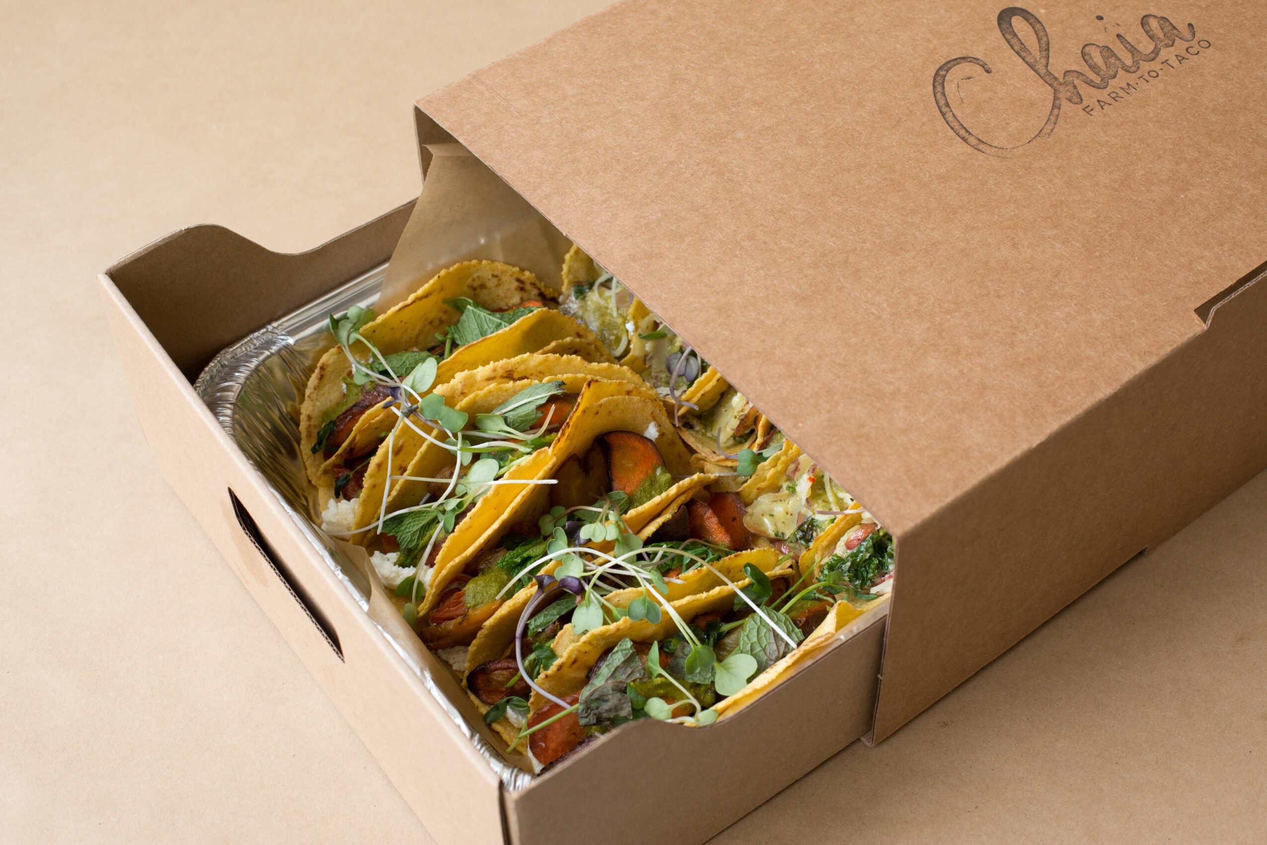 Open cardboard box containing tacos with toppings and garnishes, on a beige surface.