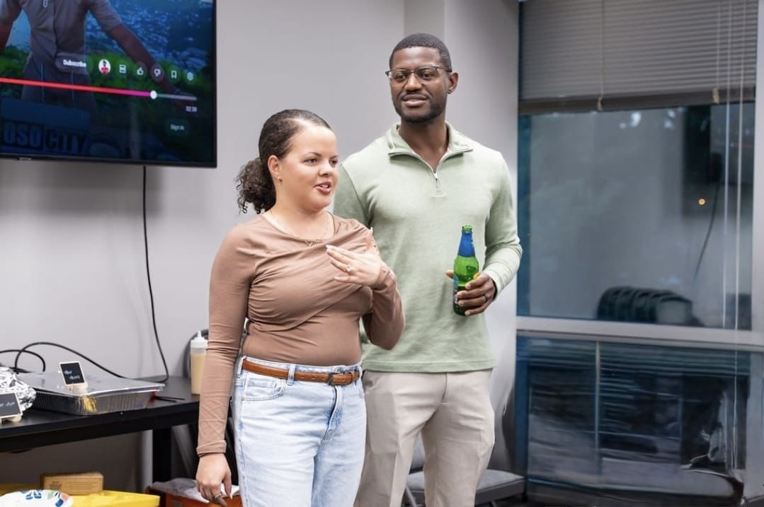 Two people standing in a room, one holding a drink, with a large window and a television screen behind them.