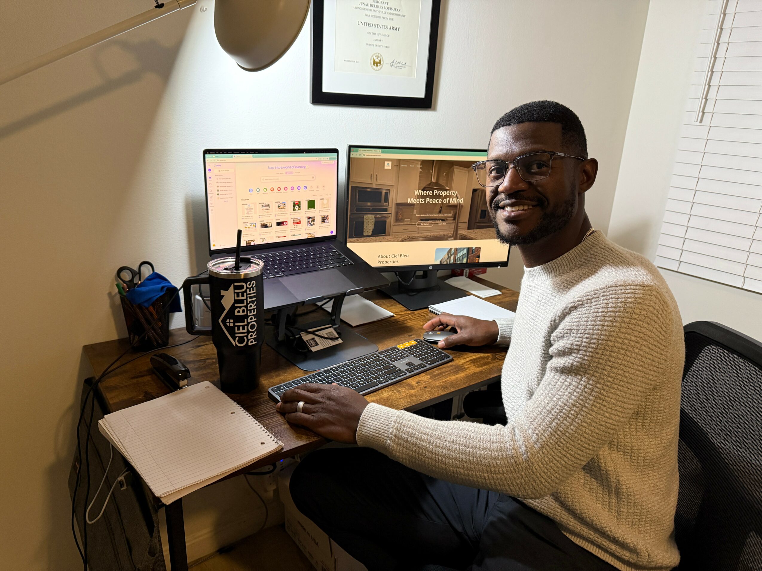 Man sitting at desk with dual monitors, smiling, wearing glasses and a white sweater, in an office setting.